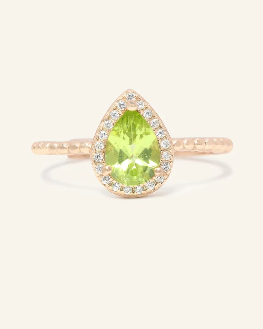 A rose gold ring featuring a vibrant green peridot stone cut in a teardrop shape, surrounded by a halo of small, sparkling cubic zirconias, with a textured band.