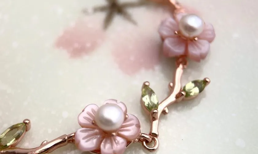 A delicate sakura twig bracelet featuring hand-carved mother-of-pearl flowers with freshwater pearl centers and peridot gemstone leaves, set in a rose gold finish. Elegant and nature-inspired.