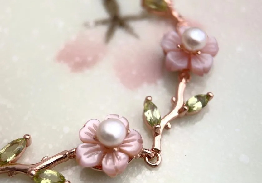 A delicate sakura twig bracelet featuring hand-carved mother-of-pearl flowers with freshwater pearl centers and peridot gemstone leaves, set in a rose gold finish. Elegant and nature-inspired.