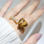 A model wearing the tiger eye heart ring. The ring features a large, polished heart-shaped tiger eye gemstone set in a yellow gold band, with the stone's characteristic golden-brown hues and chatoyancy creating a striking visual effect.