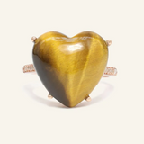A close-up image of a tiger eye heart ring. The ring features a large, polished heart-shaped tiger eye gemstone set in a rose gold band, with the stone's characteristic golden-brown hues and chatoyancy creating a striking visual effect.