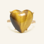 A close-up image of a tiger eye heart ring. The ring features a large, polished heart-shaped tiger eye gemstone set in a yellow gold band, with the stone's characteristic golden-brown hues and chatoyancy creating a striking visual effect.